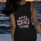 Woman wearing a black sleeveless bodycon dress with "Anti Social Social Club" graphic print on the back, standing by the sea at sunset.